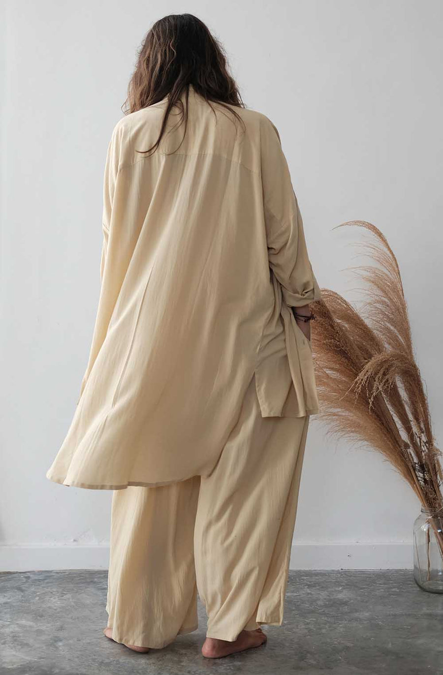 Person wearing a beige outfit standing against a white wall with dried pampas grass.