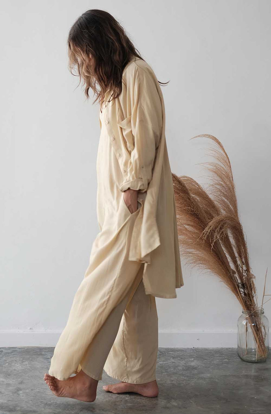 Person wearing a beige outfit standing next to a jar with dried pampas grass against a white wall.