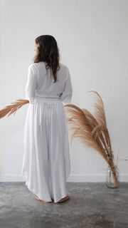 Woman in a white dress standing next to a vase with feathers on a white wall.