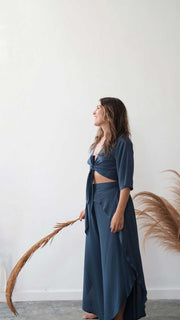 Woman in a blue outfit standing against a white wall with dried plants.