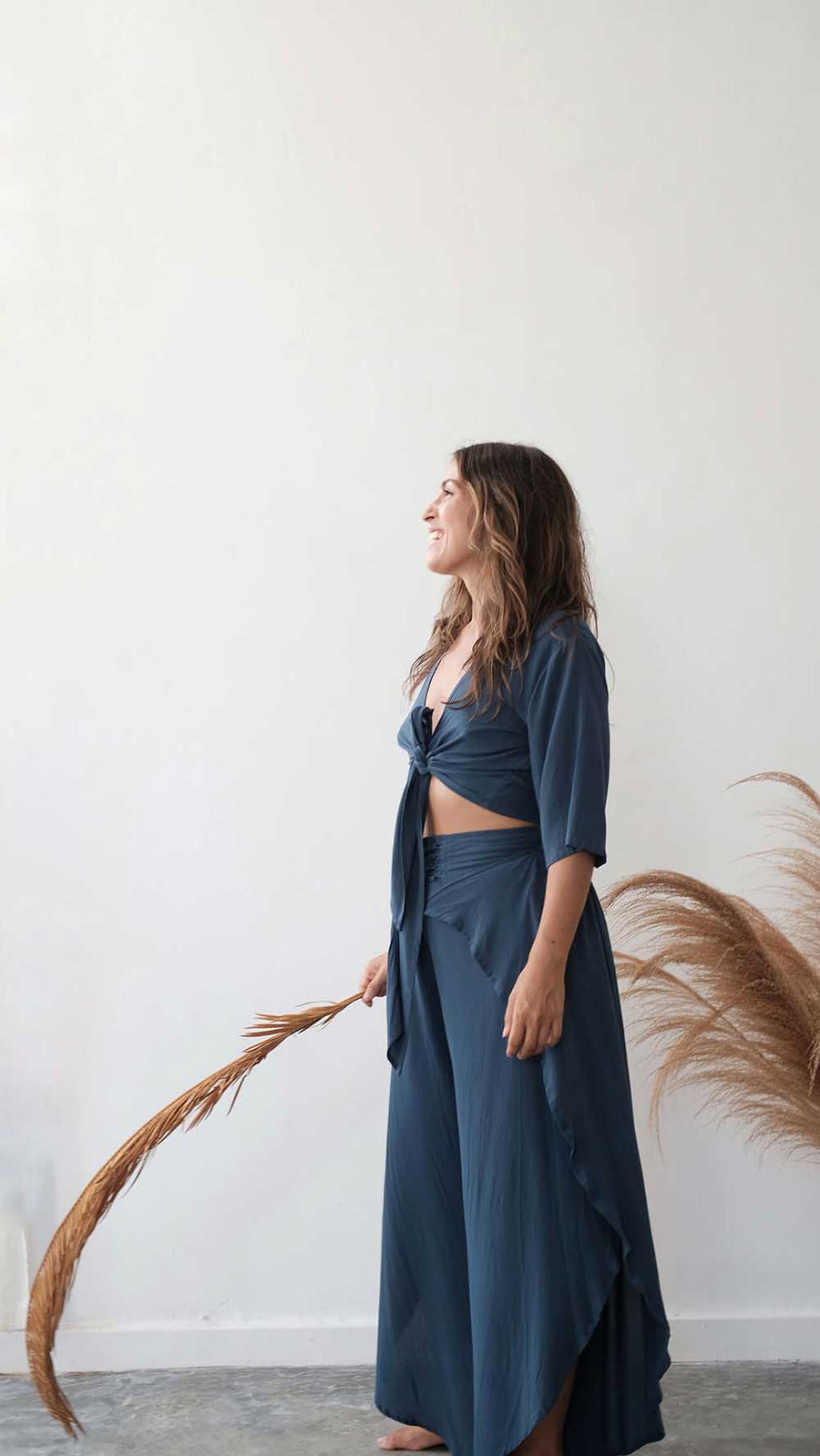 Woman in a blue outfit standing against a white wall with dried plants.
