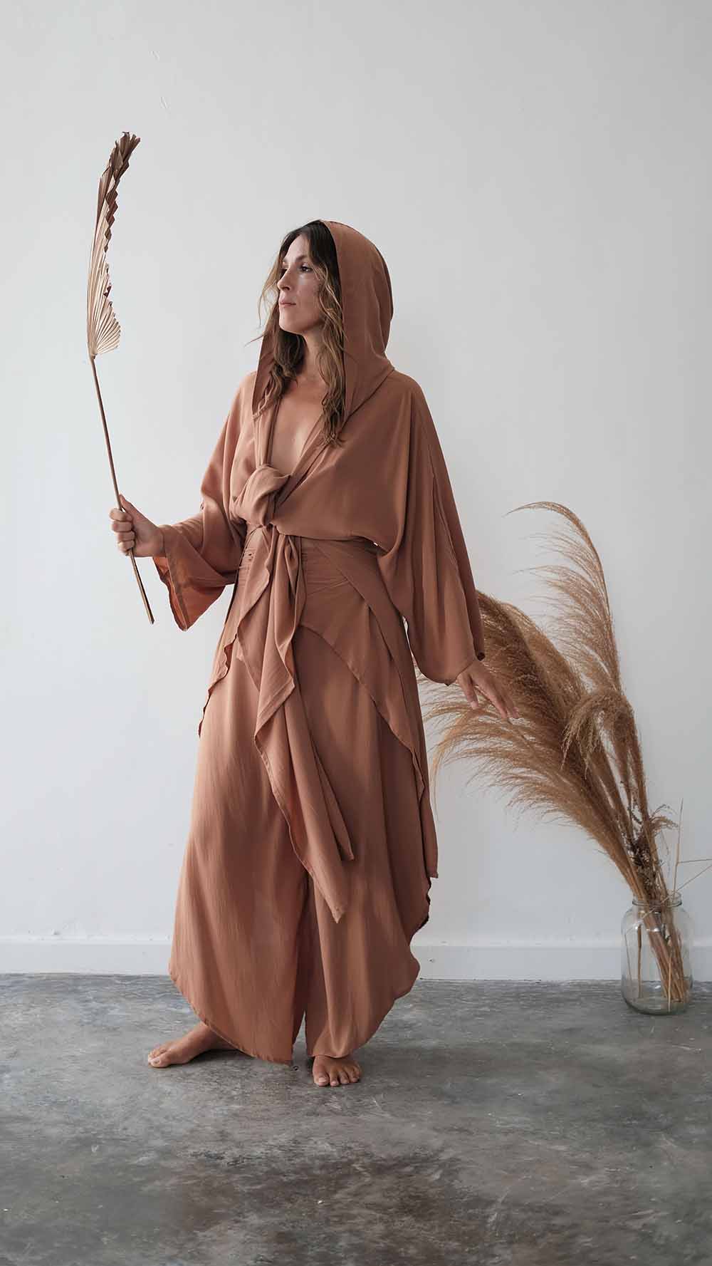Person wearing a brown hooded garment holding dried plants against a white wall.