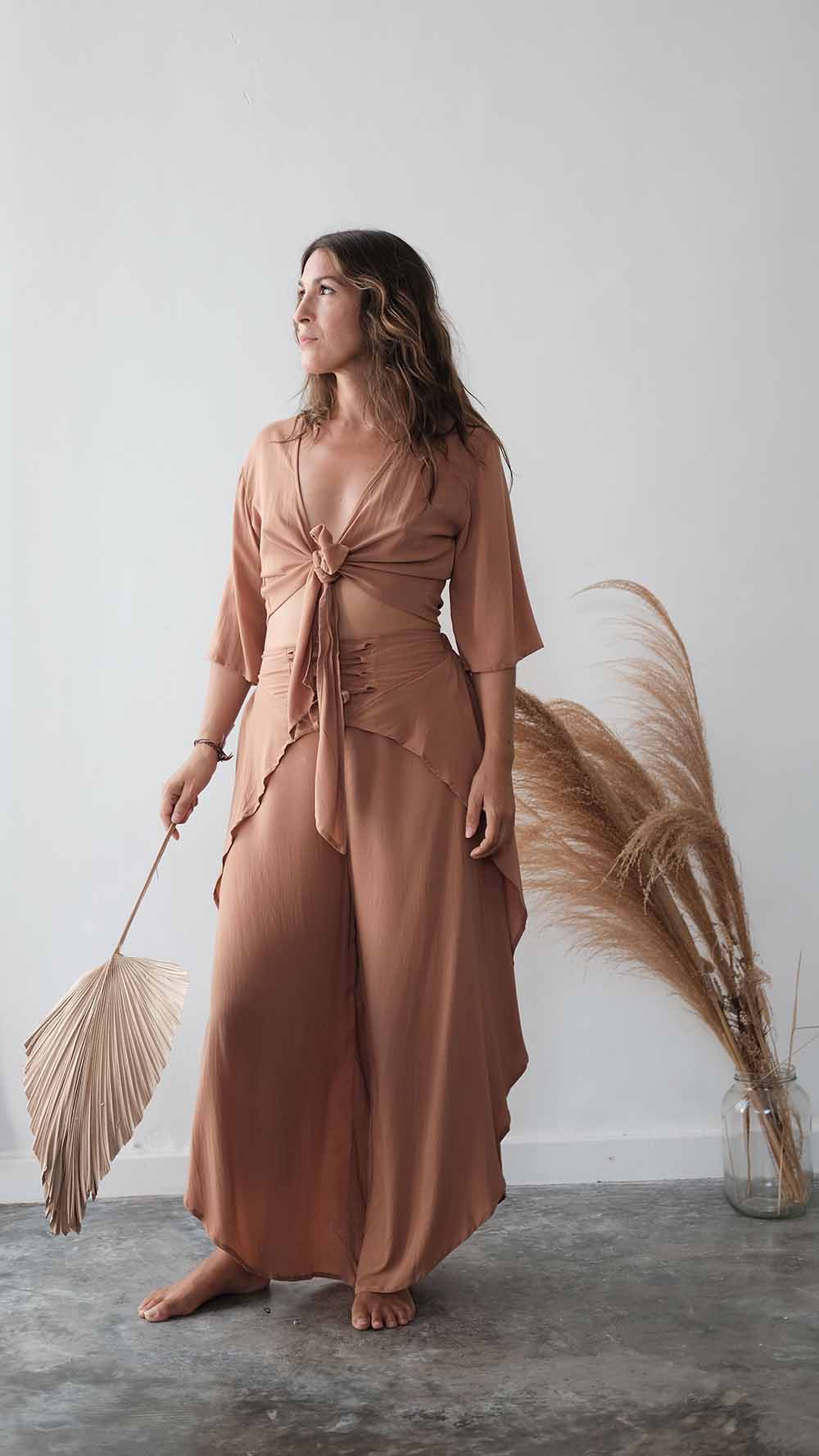 Woman in a long, light brown dress holding dried pampas grass against a white wall.