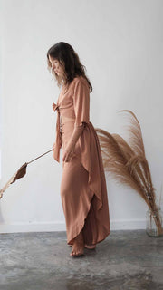 Woman in a long, light brown dress holding dried plants against a white wall.