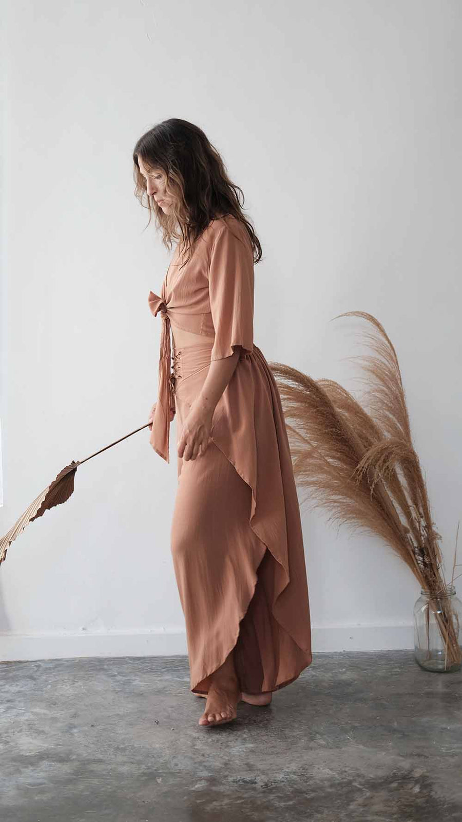 Woman in a long, light brown dress holding dried plants against a white wall.