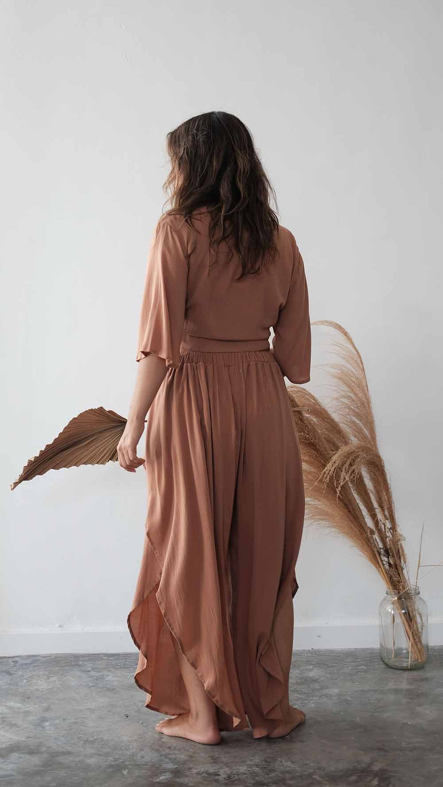 Woman wearing a brown dress standing in a minimalistic room with dried plants.