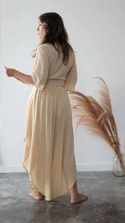 Woman wearing a beige dress standing in a minimalistic room with a vase of dried plants.