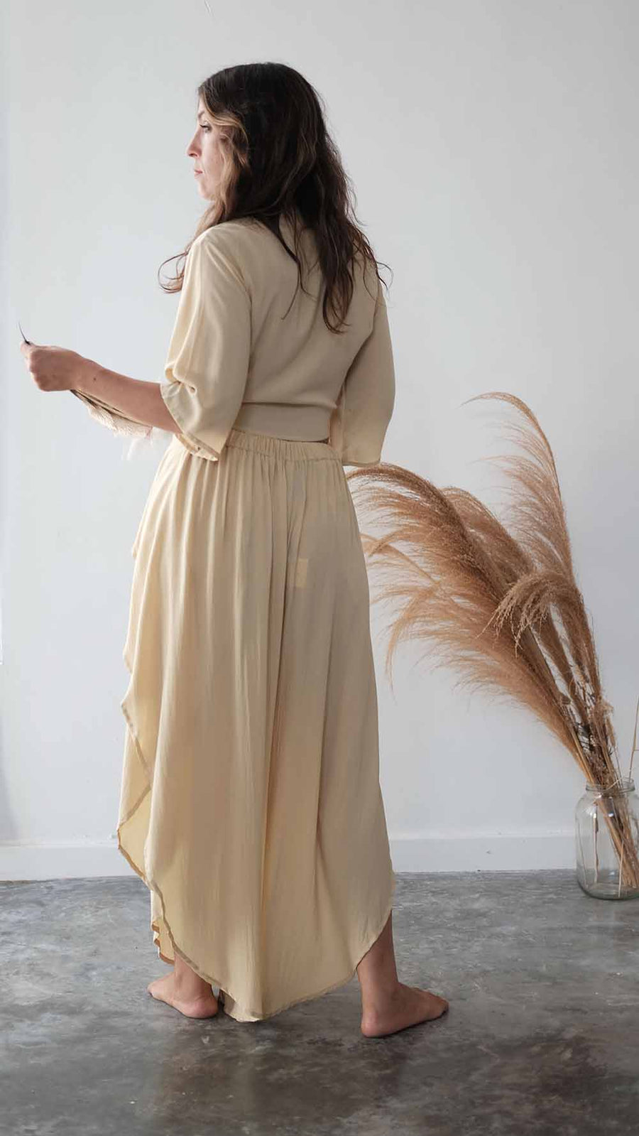 Woman wearing a beige dress standing in a minimalistic room with a vase of dried plants.