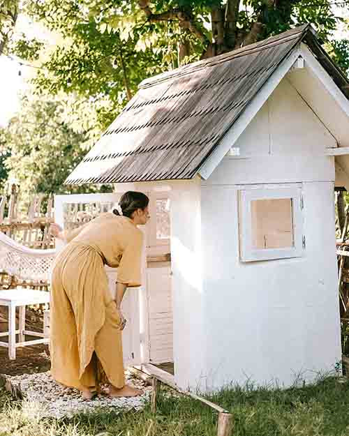 Person in a yellow dress standing next to a small white building with a thatched roof in a natural setting.