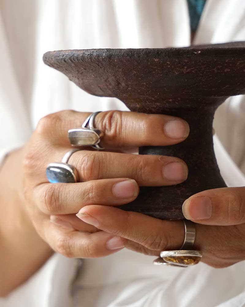 Close-up of hands holding a dark brown ceramic bowl with silver rings on fingers.