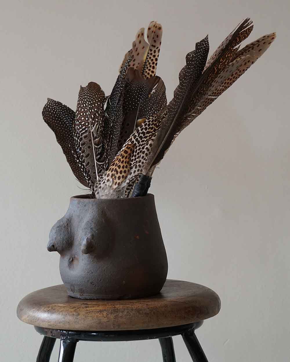 Ceramic vase with feather arrangement on a wooden stool against a plain background