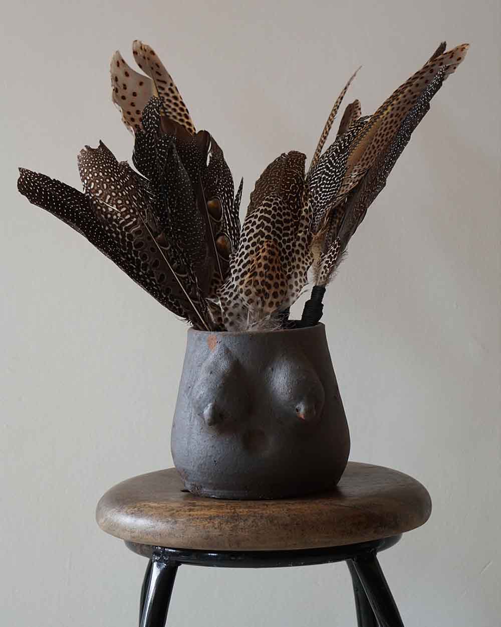Black vase shaped like a female torso with feathers on a wooden stool against a plain background