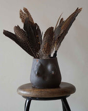 Black vase shaped like a female torso with feathers on a wooden stool against a plain background