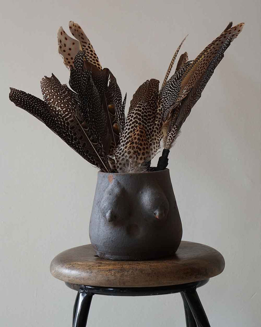 Black vase shaped like a female torso with feathers on a wooden stool against a plain background