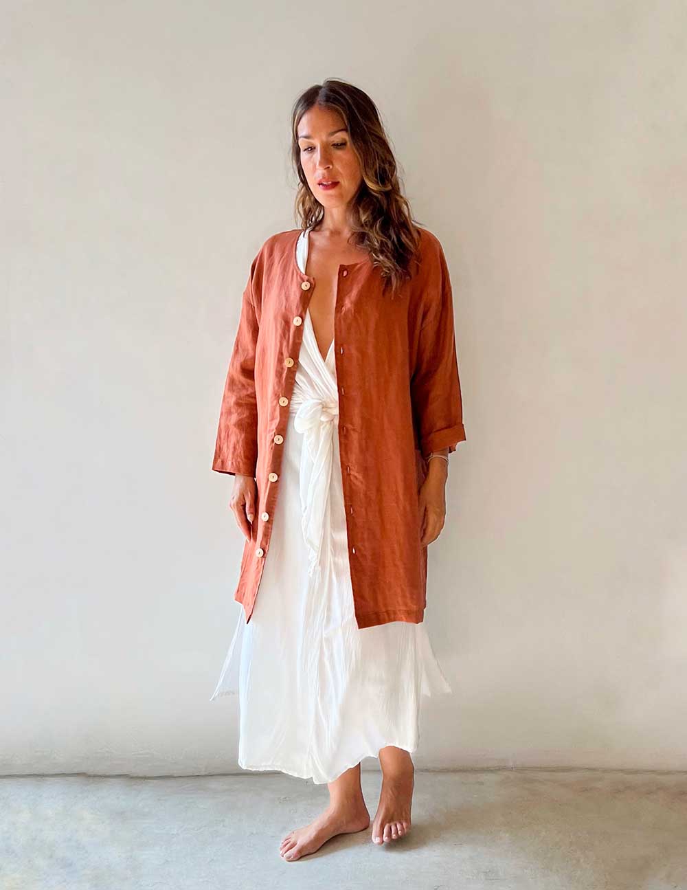 Woman wearing a rust-colored jacket over a white dress against a plain background