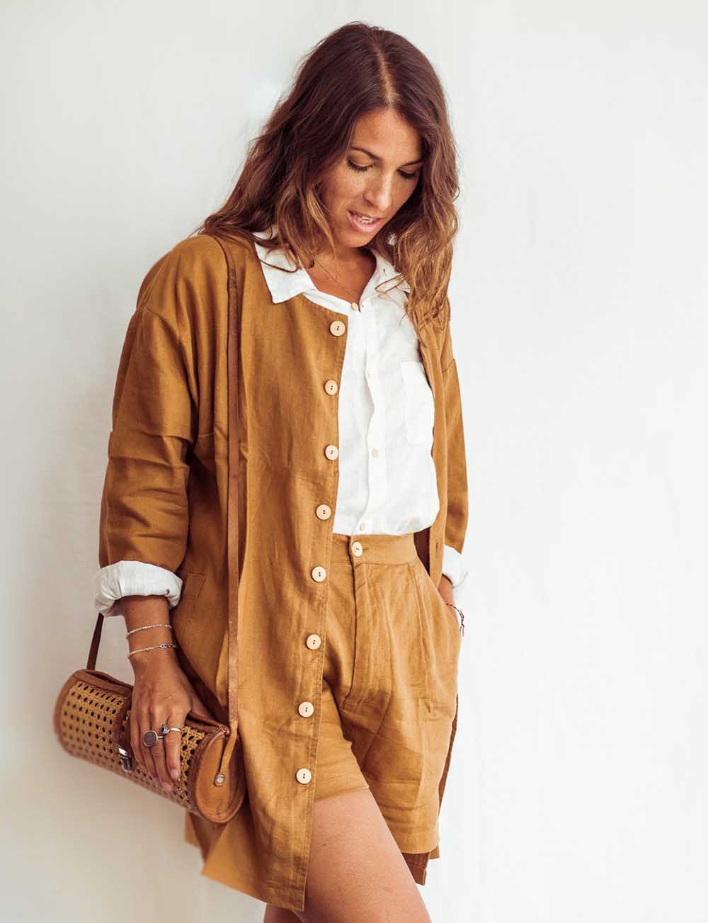 Woman wearing a brown dress with a white shirt underneath, holding a woven handbag against a white background.