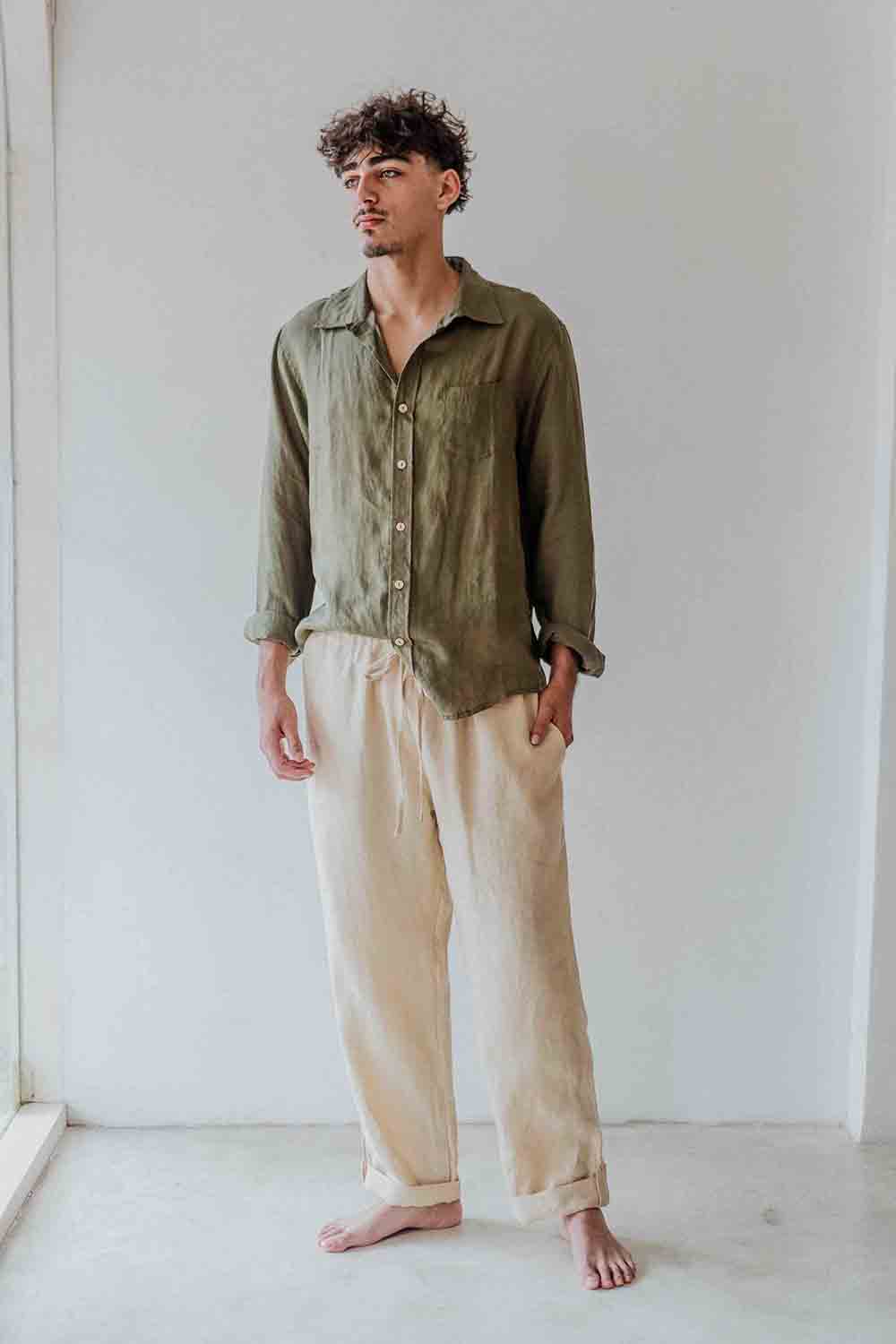 Man wearing an olive green shirt and beige pants standing against a white wall.