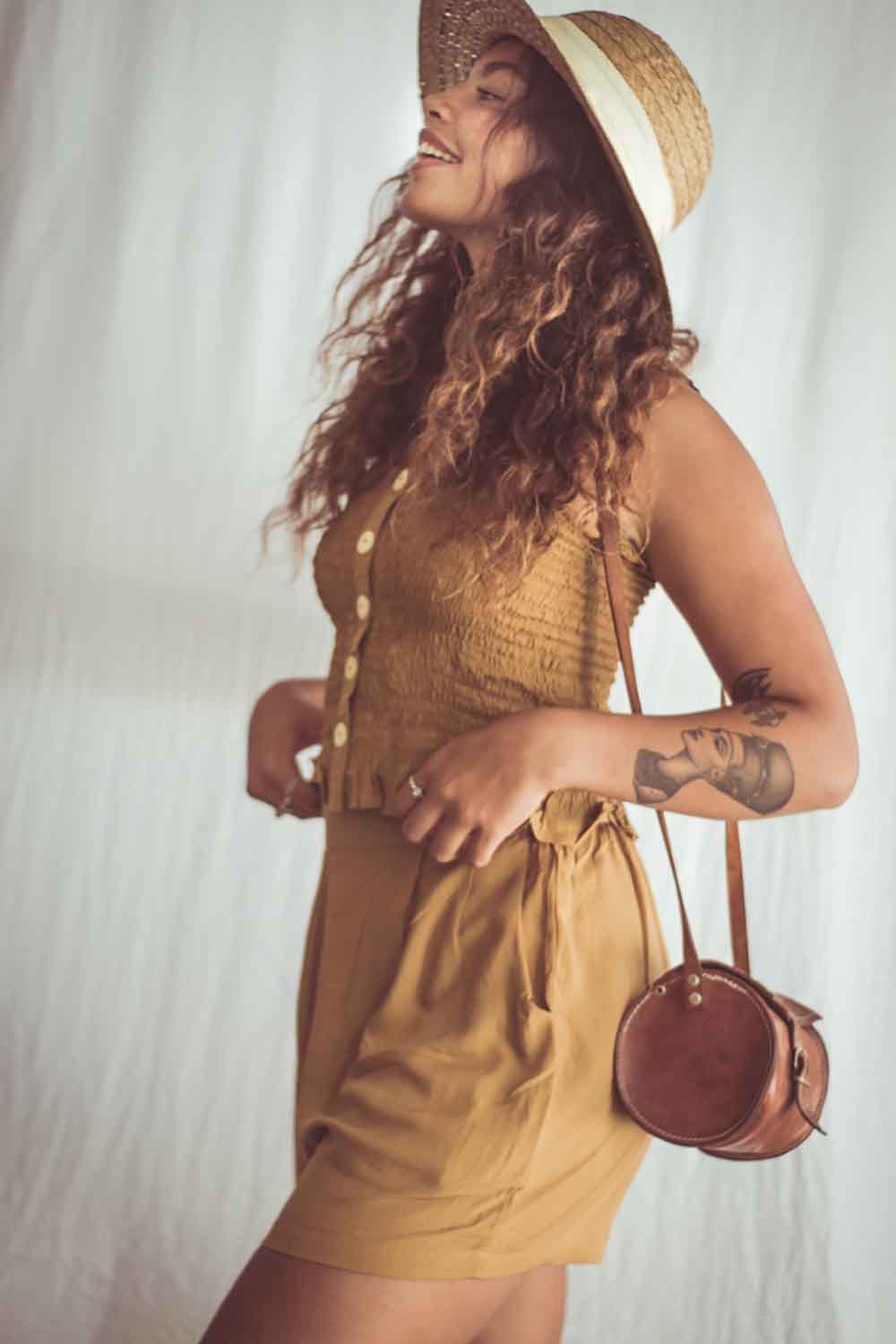 Woman wearing a mustard yellow dress and hat, holding a brown leather bag against a white background