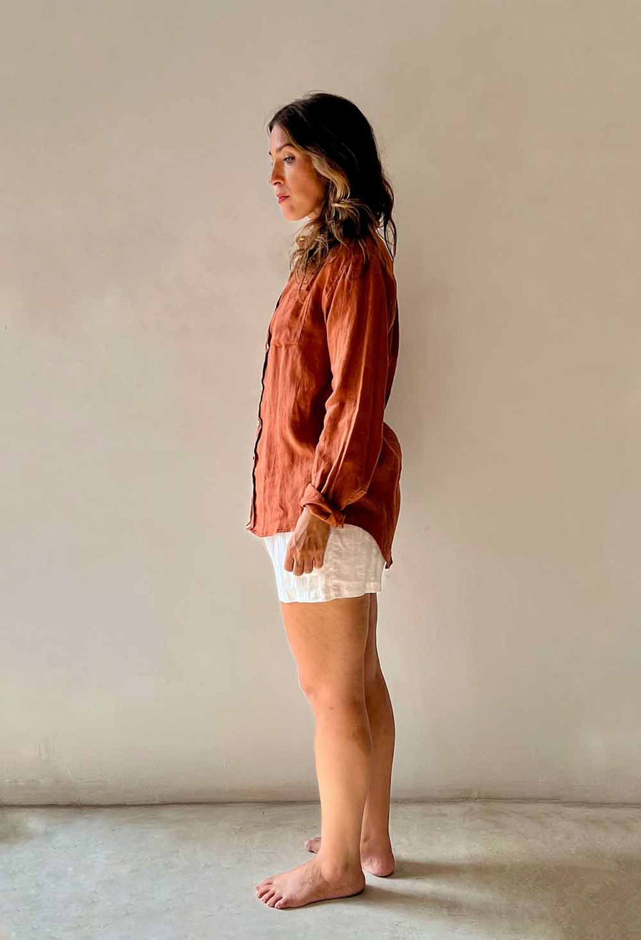 Woman wearing a rust-colored shirt and white shorts standing against a plain wall.
