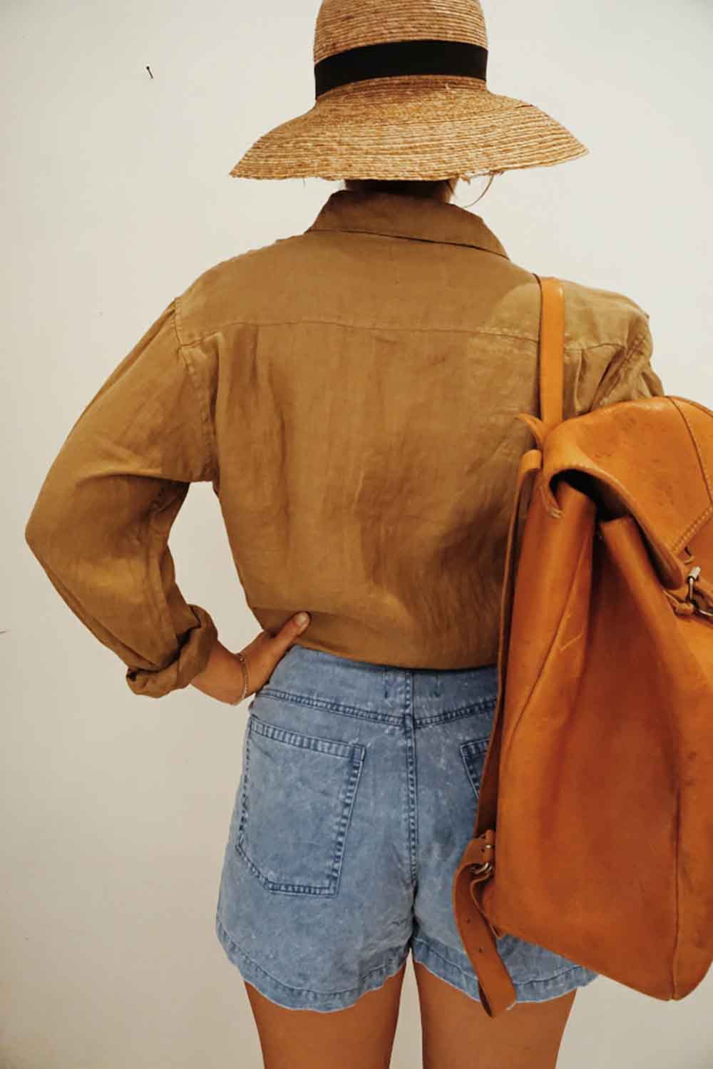 Person wearing a brown jacket, blue shorts, and a straw hat with an orange backpack on a plain background