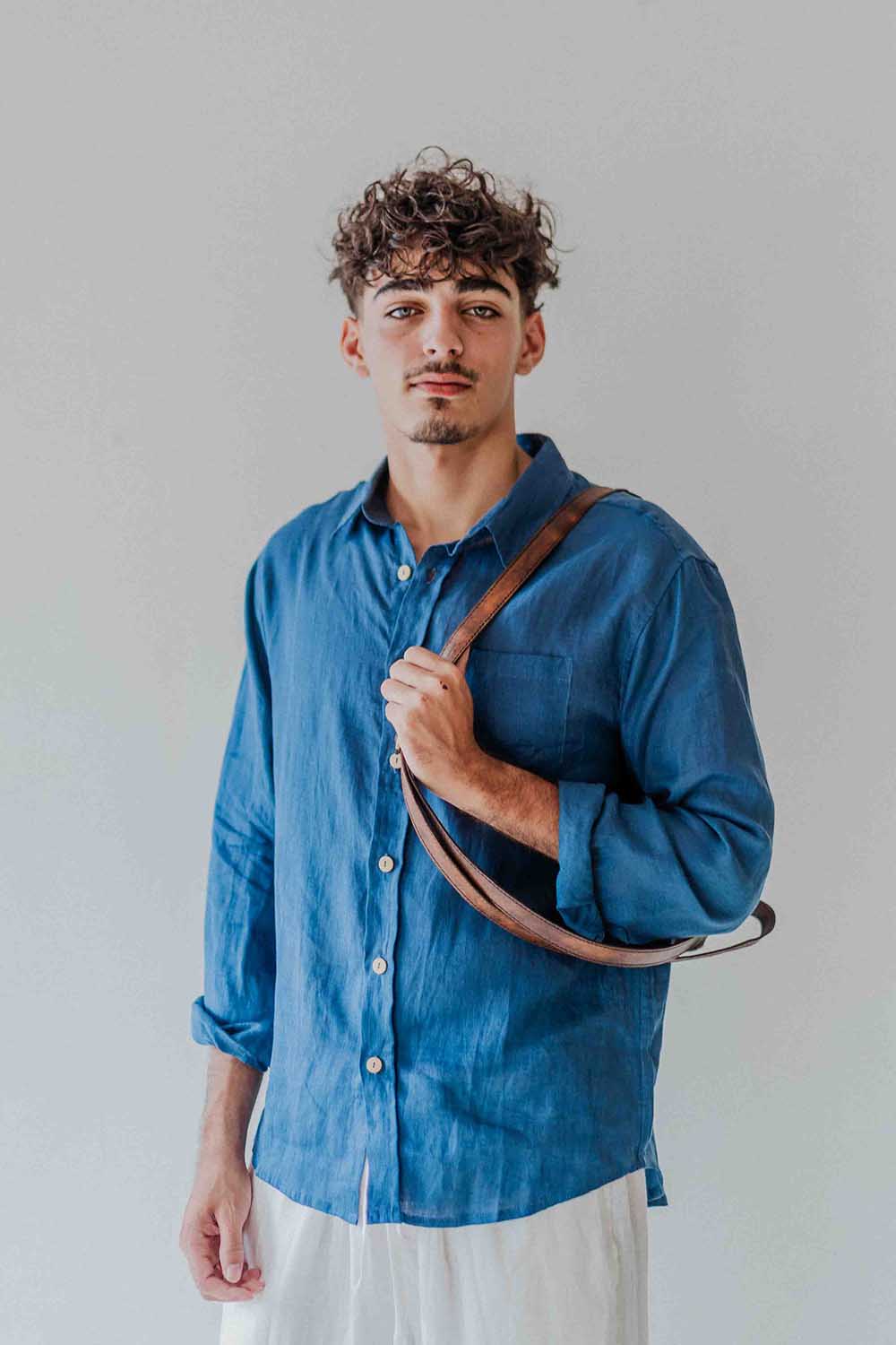 Man wearing a blue shirt holding a brown leather bag against a plain background