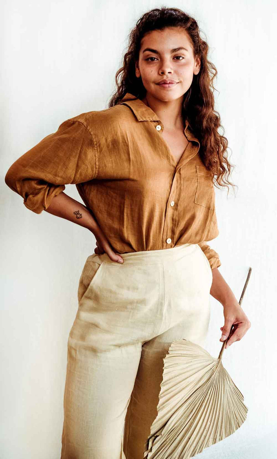 Woman wearing a mustard yellow shirt and beige skirt holding a fan against a white background