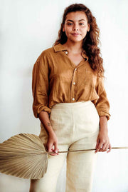 Woman wearing a mustard yellow shirt and beige pants holding a dried palm leaf against a white background