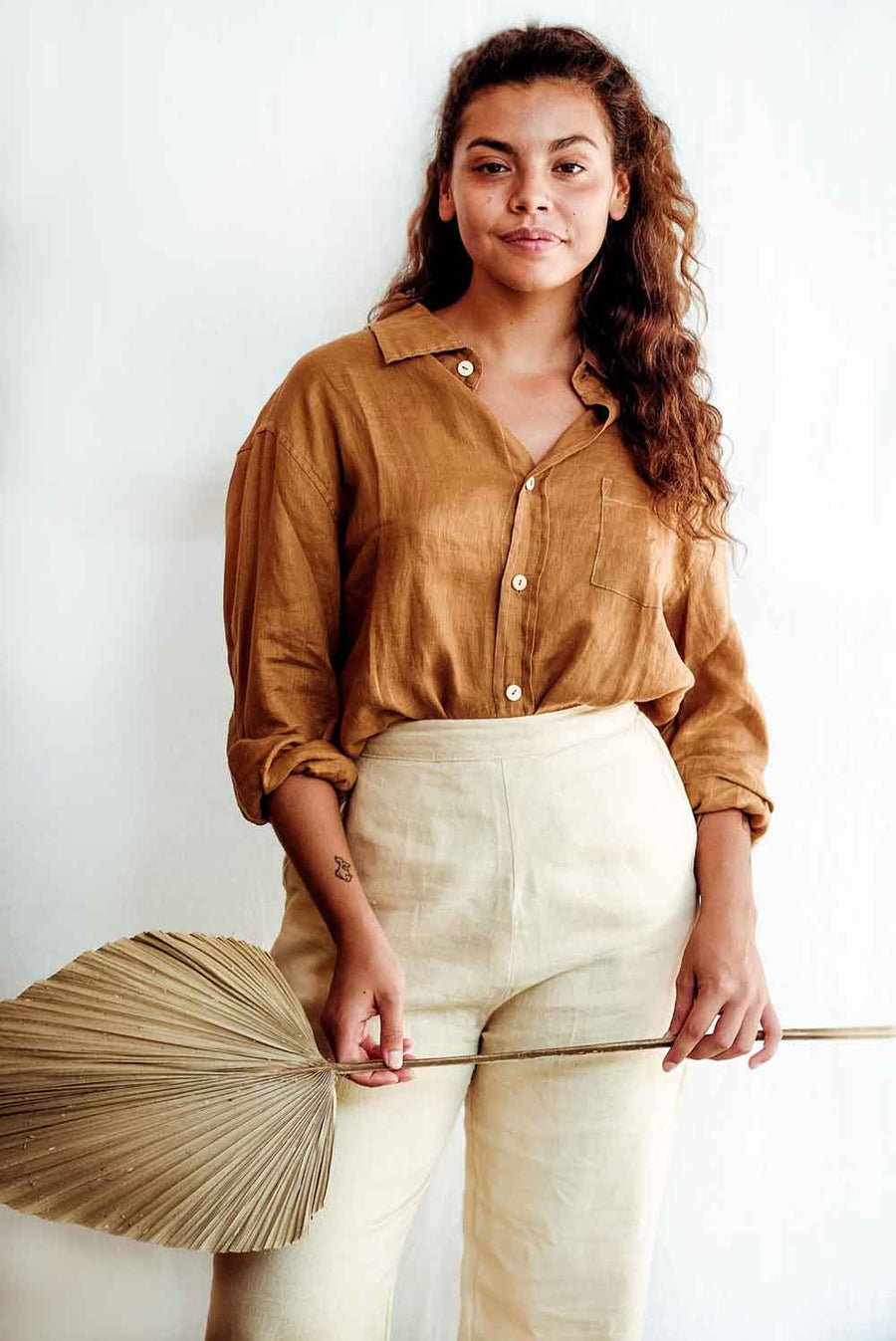 Woman wearing a mustard yellow shirt and beige pants holding a dried palm leaf against a white background