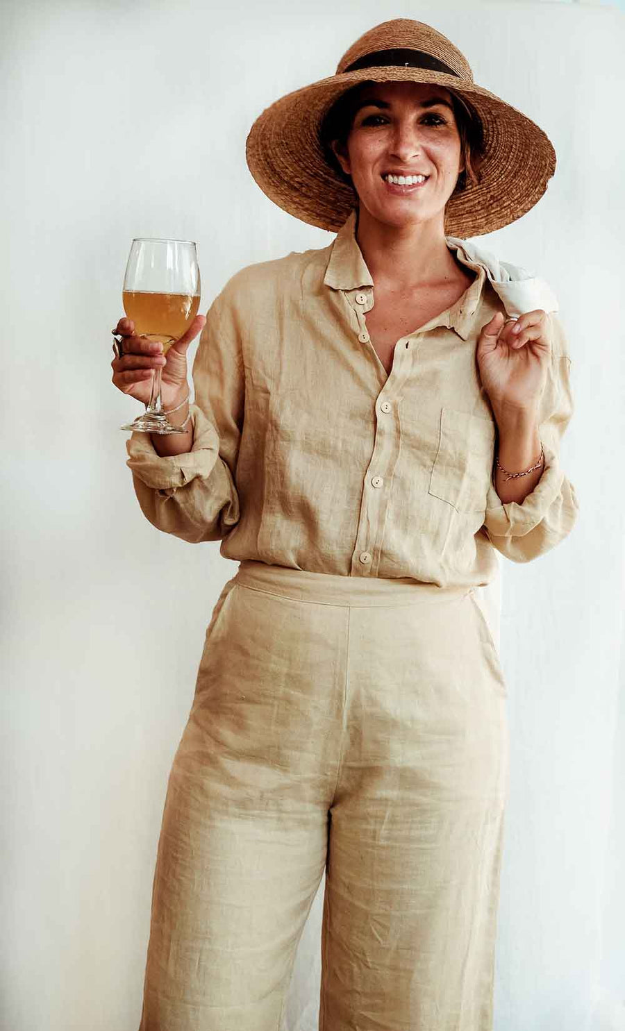 Woman in beige linen outfit holding a glass of wine against a white background