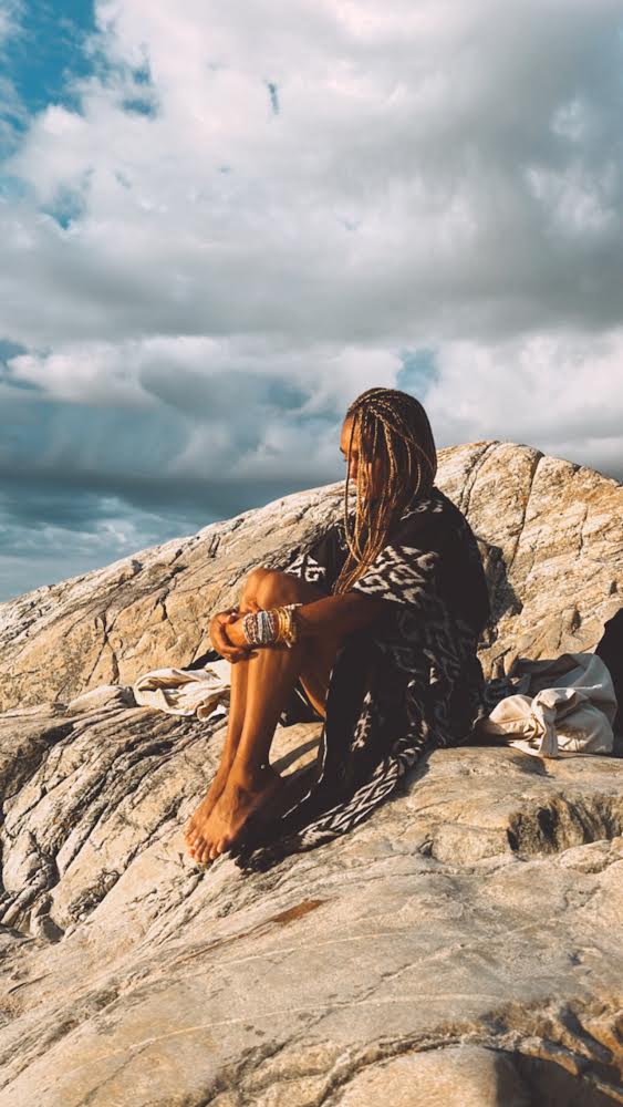 A person sitting on rocky terrain wearing a black kaftan with a traditional Ikat pattern, paired with a matching belt.