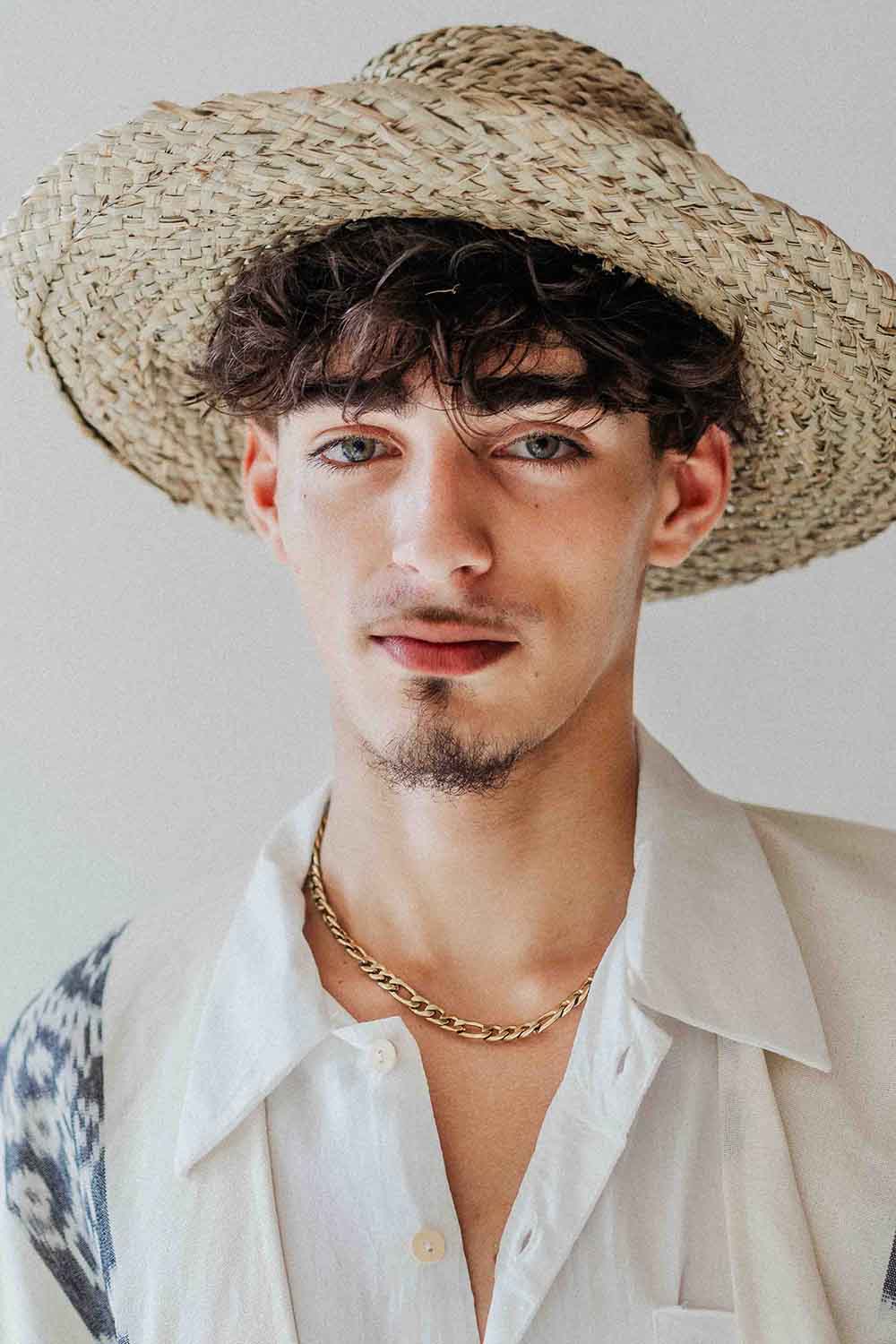 Man wearing a straw hat and white shirt with a gold chain necklace against a plain background