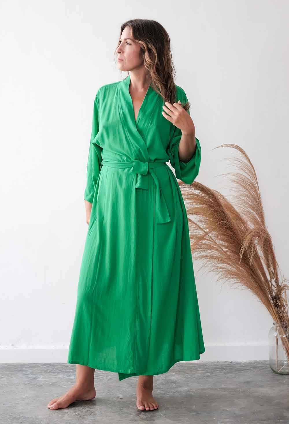 Woman wearing a green dress standing next to dried pampas grass against a white wall.