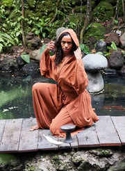 Person in a long, orange robe sitting on a wooden platform by a pond with greenery in the background