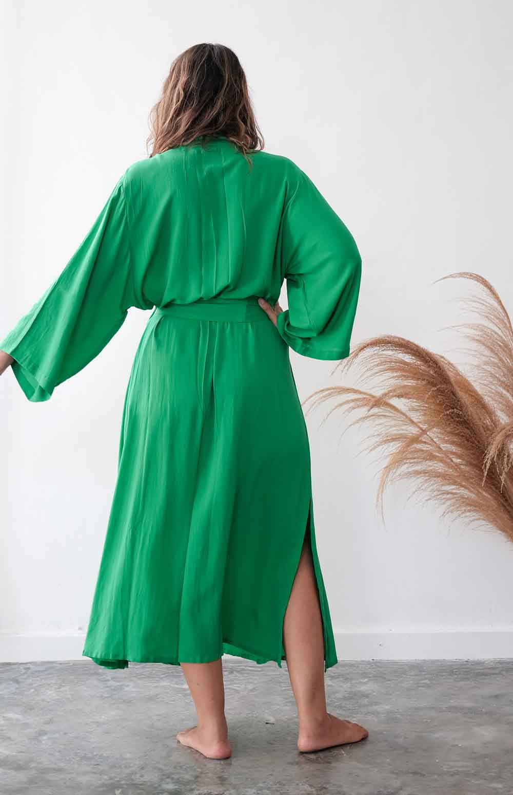 Person wearing a green robe standing against a white wall with dried plants.