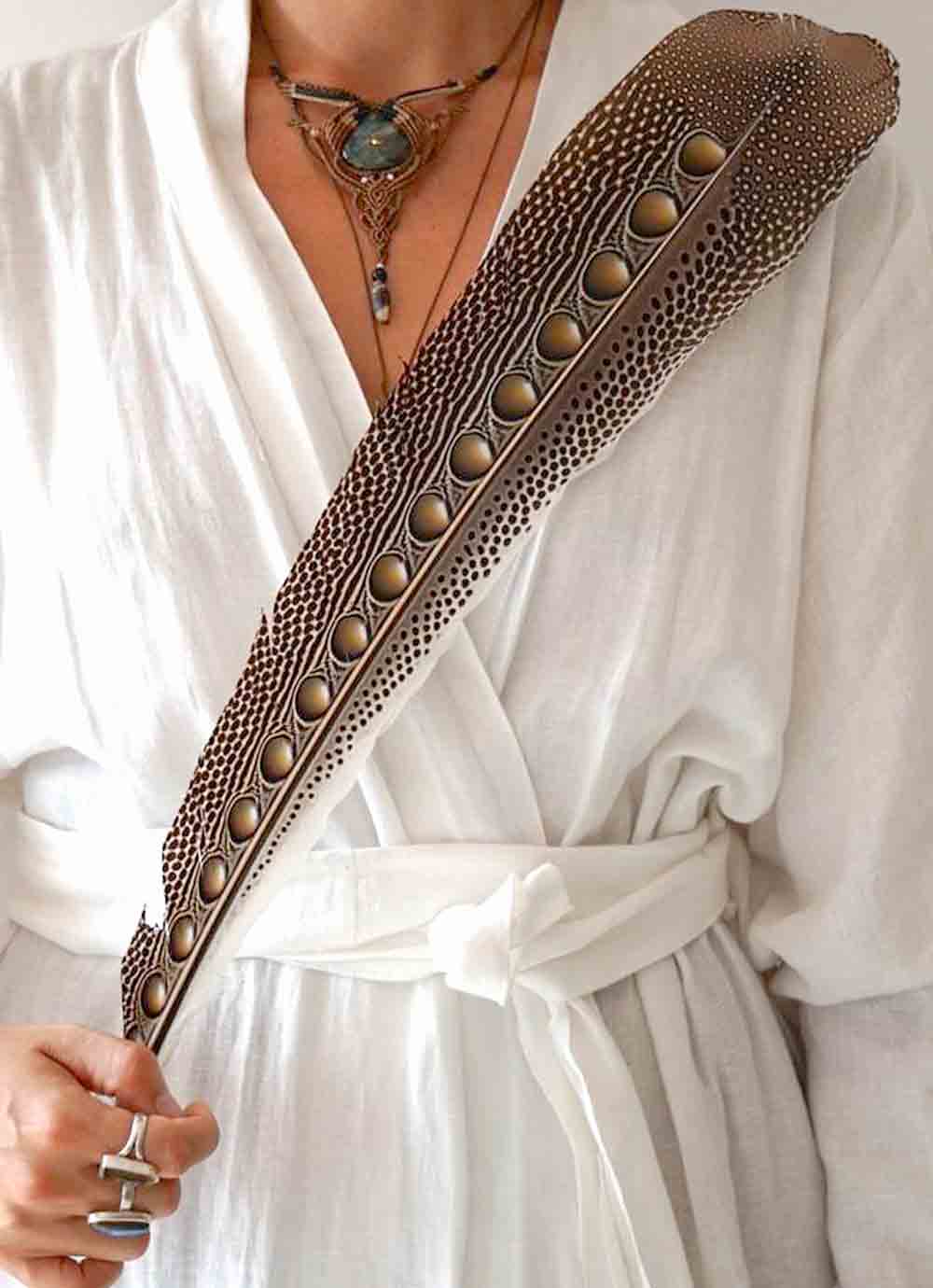 Person holding a decorative feathered staff with intricate designs, wearing a white robe.