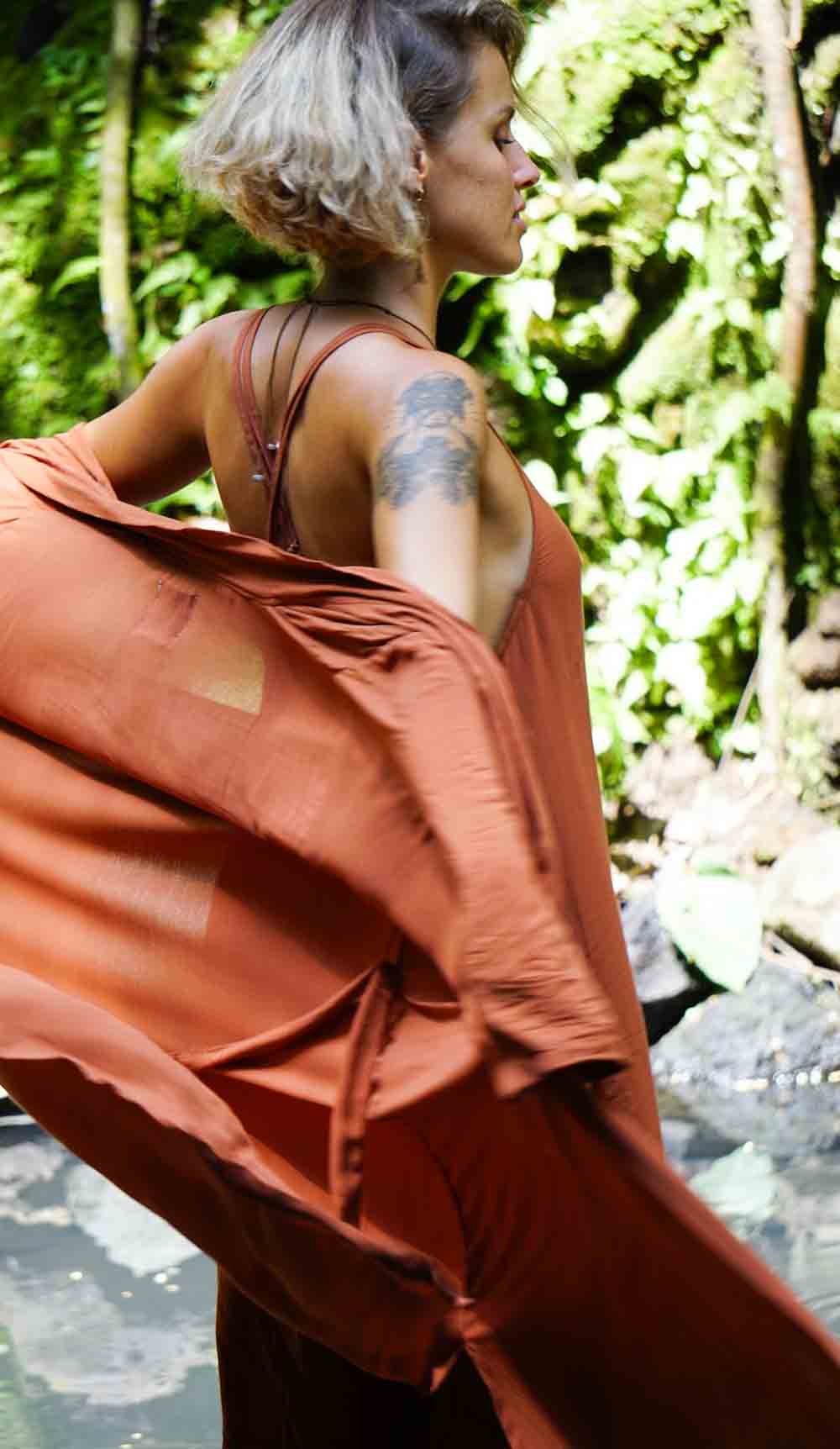 Person wearing a rust-colored outfit standing in a natural setting with greenery.