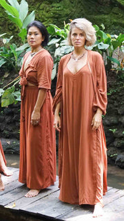 Two women in rust-colored long dresses standing on a wooden platform with greenery in the background.