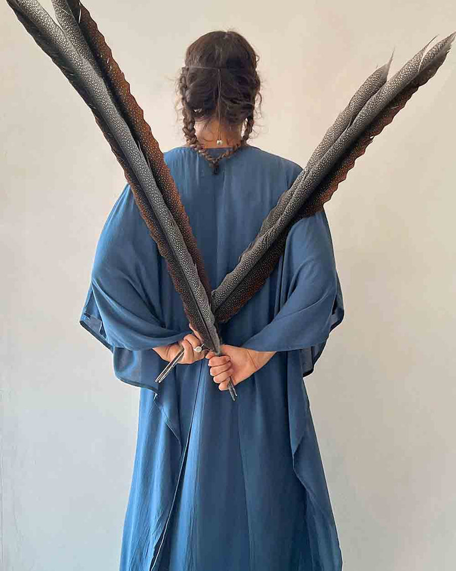 Person in a blue robe holding two large feathers against a plain background
