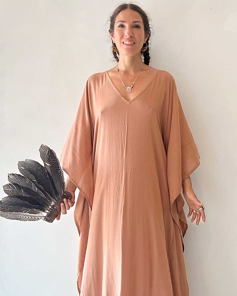 Woman wearing a long, light brown dress holding a peacock feather against a plain background