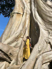 Person in a yellow dress standing next to a large tree with intricate bark patterns