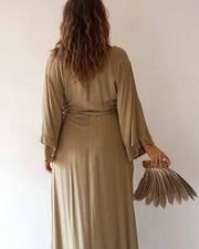 Woman in a long beige dress holding a matching clutch against a plain background