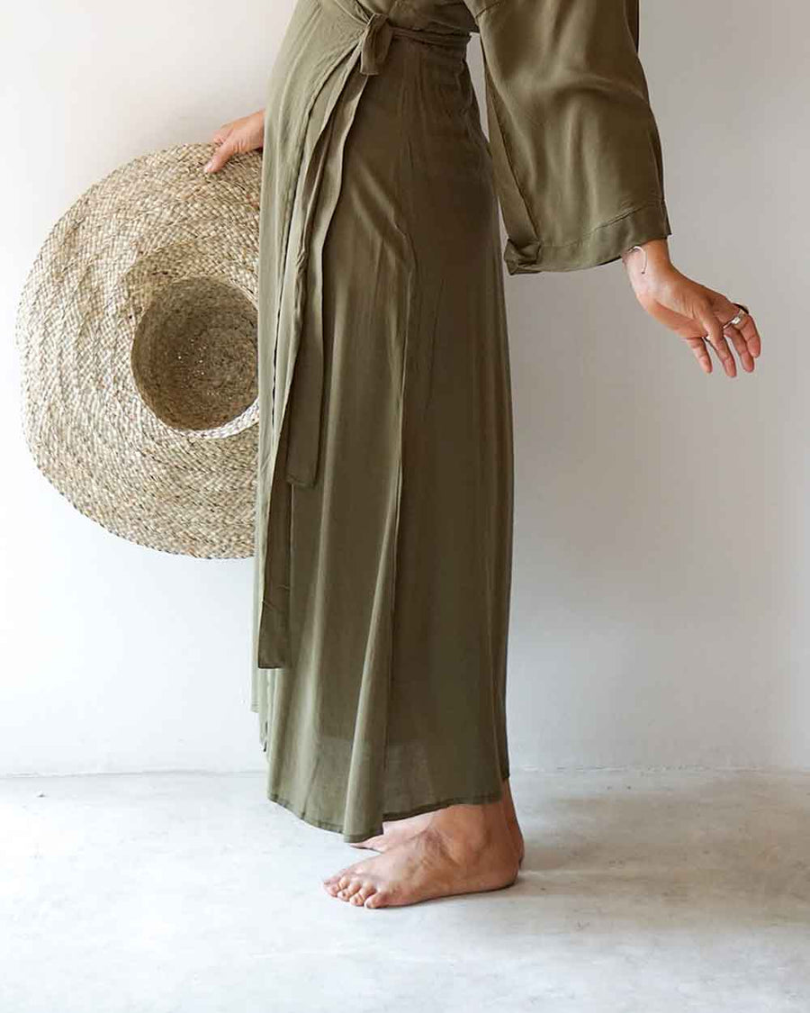 Person wearing a long olive green dress holding a straw hat on a white background