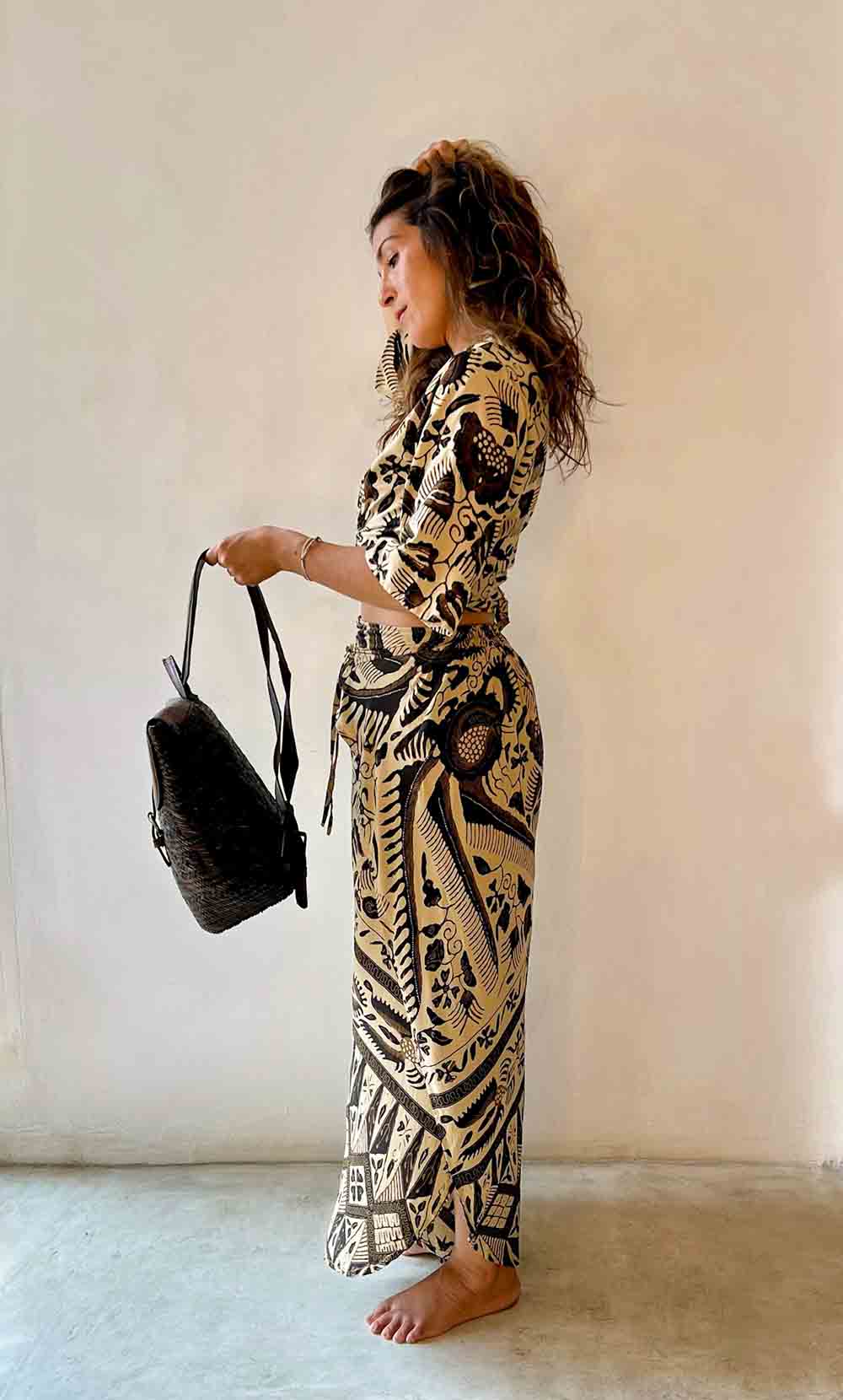Woman in a black and gold patterned outfit holding a black bag against a plain background