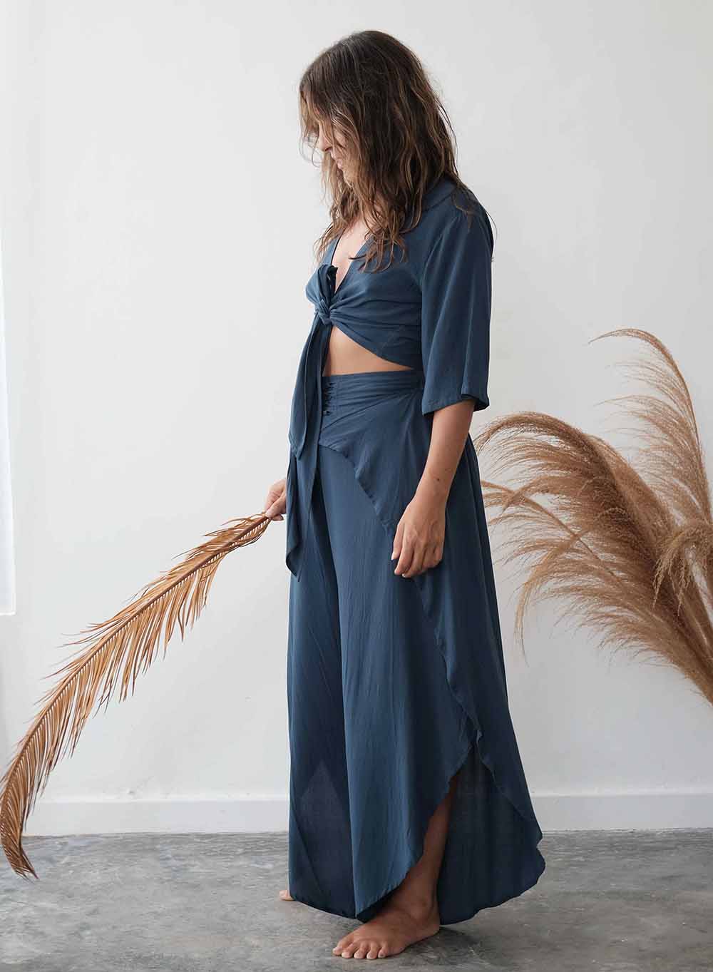 Woman wearing a blue outfit holding a dried plant against a white wall.