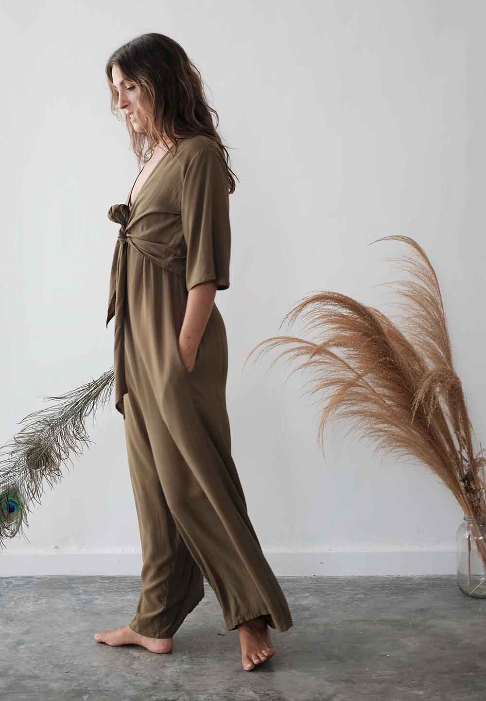 Woman wearing a brown jumpsuit standing against a white wall with decorative plants.