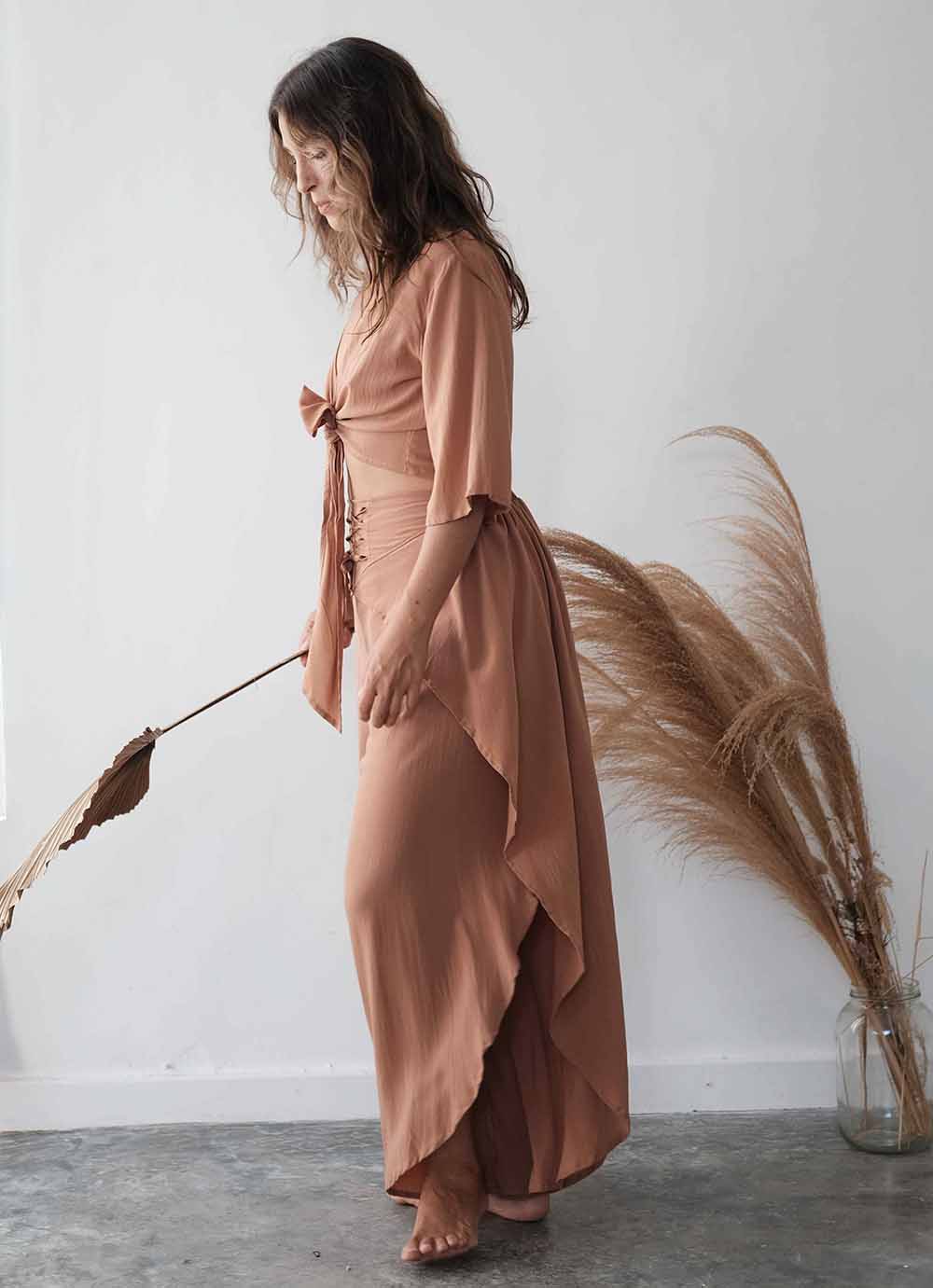 Woman in a long, flowing dress standing next to dried pampas grass against a white wall.
