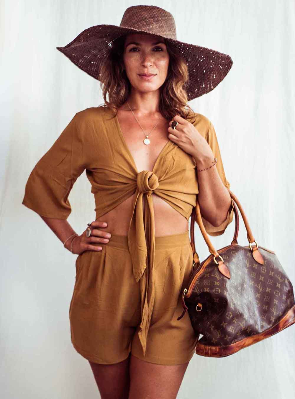 Woman in a mustard yellow outfit with a wide-brimmed hat and brown handbag against a white background