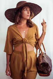 Woman in a mustard yellow outfit with a wide-brimmed hat and brown handbag against a white background