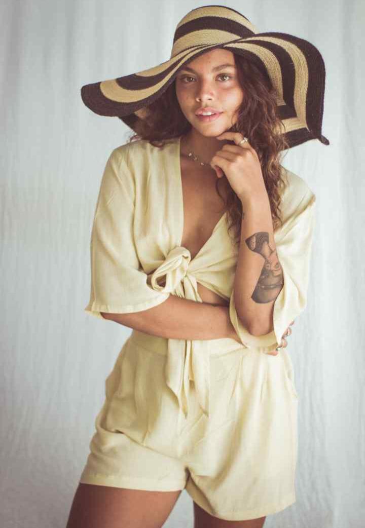 A model wearing a cropped top and shorts, with the top in a sand color, tied at the front, and the model is accessorized with a wide-brimmed hat and a necklace.