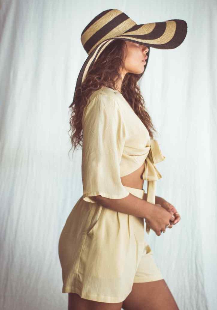 A model wearing a cropped top and shorts, with the top in a sand color, tied at the front, and the model is accessorized with a wide-brimmed hat and a necklace.